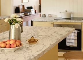 A warm yellow Kaboodle kitchen featuring honey-yellow cabinetry and white marble-look benchtops. The kitchen includes an island with a fruit bowl, a juicer, and a bar stool