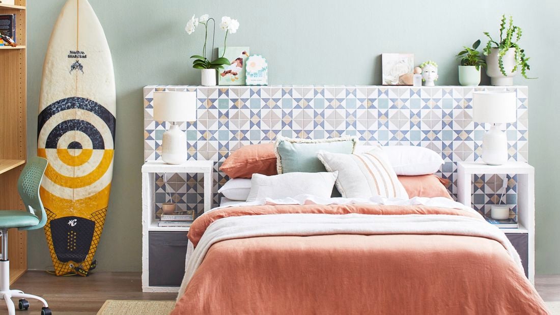  A light-coloured tiled D.I.Y. bedhead with green plants on top, behind a bed in a room with a surfboard, chair and rug