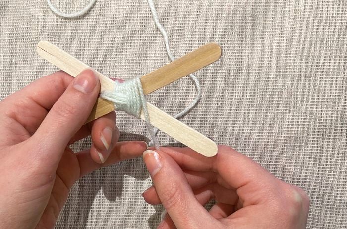 Two hands hold a white piece of yarn and two paddle pop sticks crossed over.