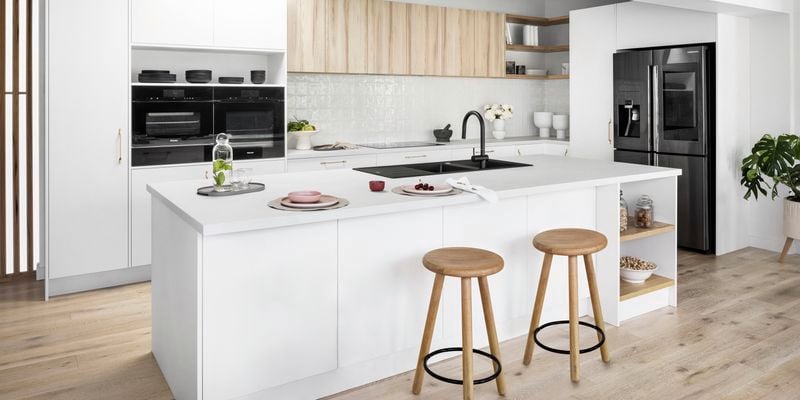 Modern white kitchen with timber bar stools and  timber feature cabinets, black sink and mixer tap and a large double door fridge.