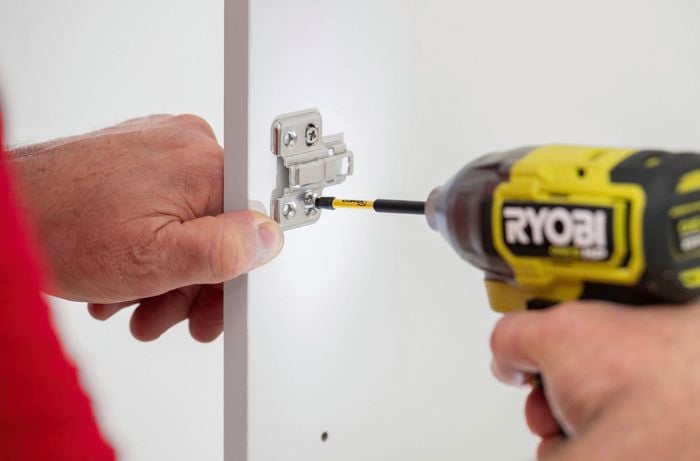 A person is using a power drill to separate the hinge arms from the mounting clip.