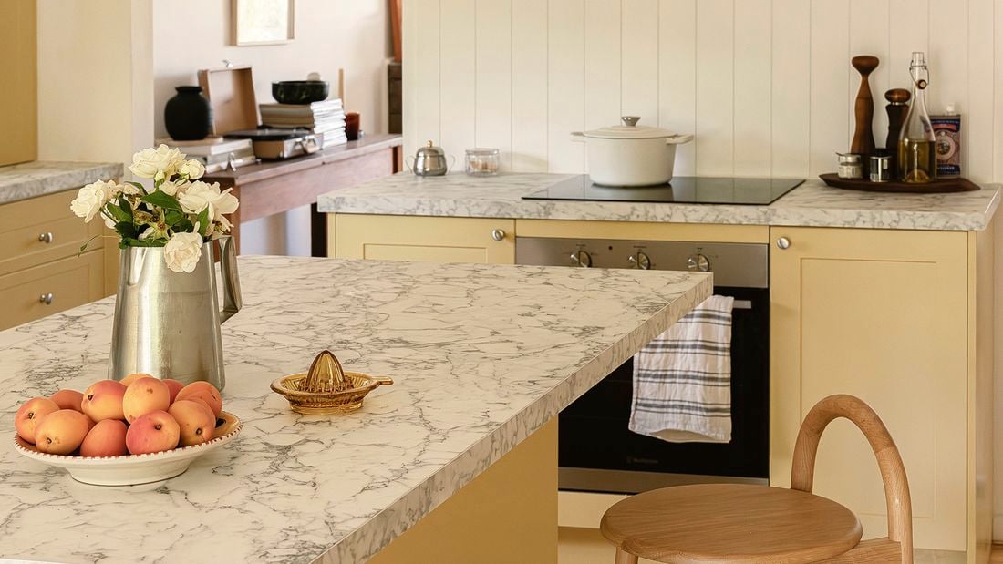 A warm yellow Kaboodle kitchen featuring honey-yellow cabinetry and white marble-look benchtops. The kitchen includes an island with a fruit bowl, a juicer, and a bar stool