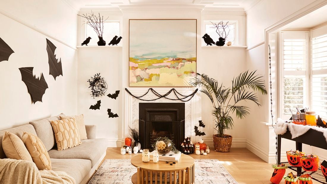 A living room decorated for Halloween with black bat cut-outs on the walls, pumpkins, candles and a table with orange jack-o'-lantern buckets. A sofa, fireplace and a large painting are also visible