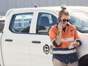 An image of a female tradie, shot from the waist up, wearing orange hi-vis and black sunglasses, is holding and looking at a piece of paper while on a mobile phone. She is standing next to a white truck