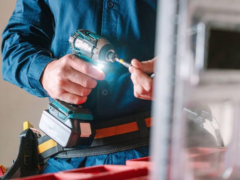 Person wearing a blue Citeco work shirt and tool belt using a cordless drill with a glowing LED light while preparing a screw bit indoors.