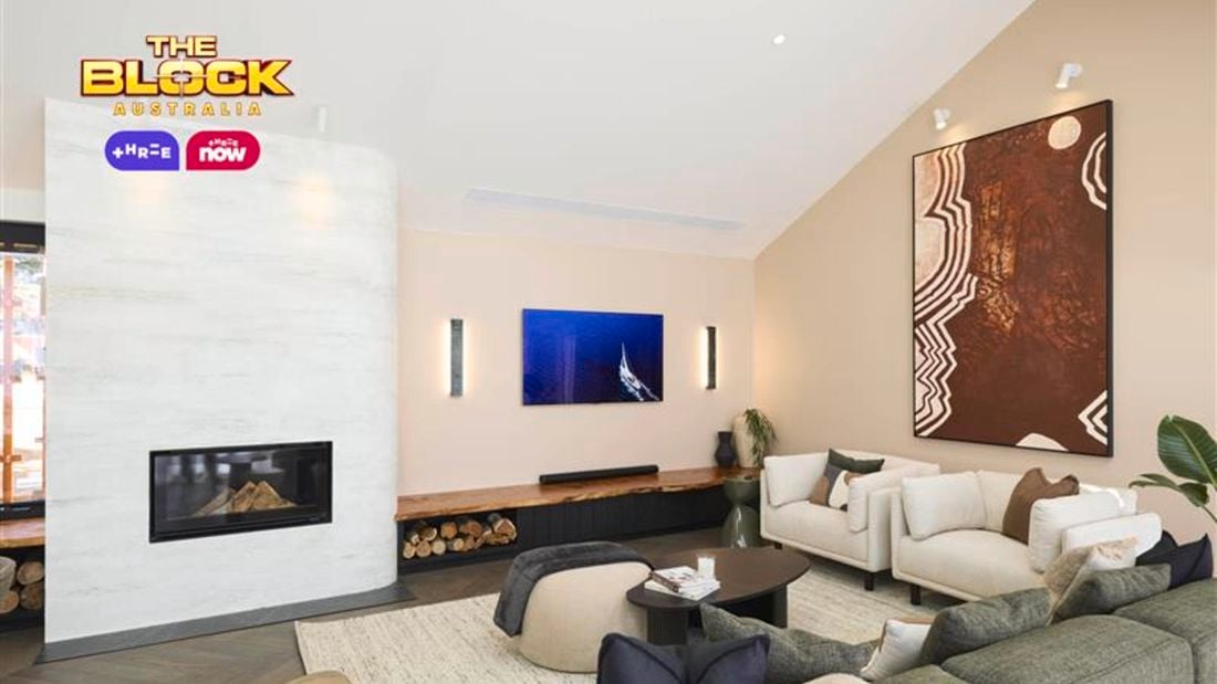 A modern living room with a high ceiling, featuring a fireplace on the left with firewood stored beneath. The space includes a green sofa, white armchairs, and a small round coffee table with books. Two large pieces of wall art are displayed—one above the fireplace and another on the right wall.