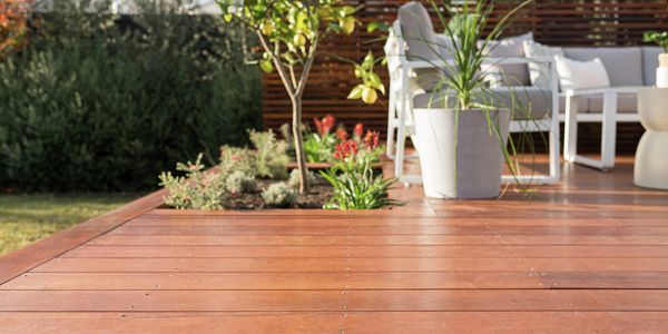 A sunlit outdoor deck with timber boards, potted plants, and outdoor seating surrounded by garden greenery.