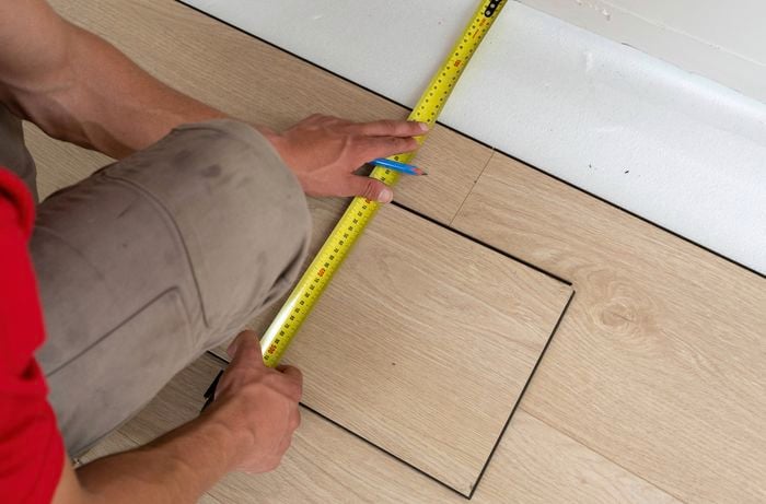 A person is measuring the distance from the edge of the last board to the wall using a tape measure to ensure proper gap coverage near the wall