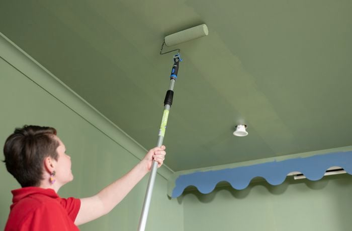 In a red T-shirt, a woman is using a paint roller to apply green paint to the ceiling in a room with green walls