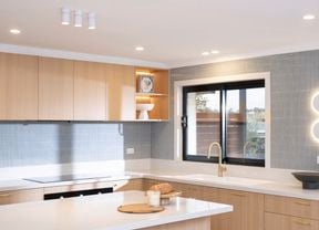 A modern kitchen features light wood cabinetry, a sleek white benchtop, vertical tile splashback, and stylish lighting.