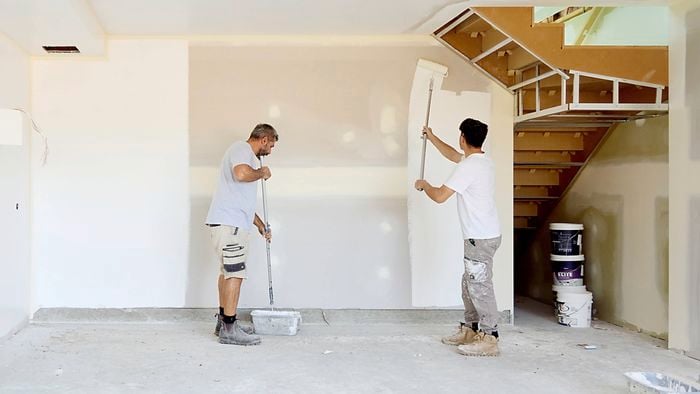 Two people painting interior walls of an unfinished room, using long-handled rollers. The space has bare concrete flooring, partially painted white walls, exposed sections around windows, and a wooden staircase with construction materials and paint buckets nearby.