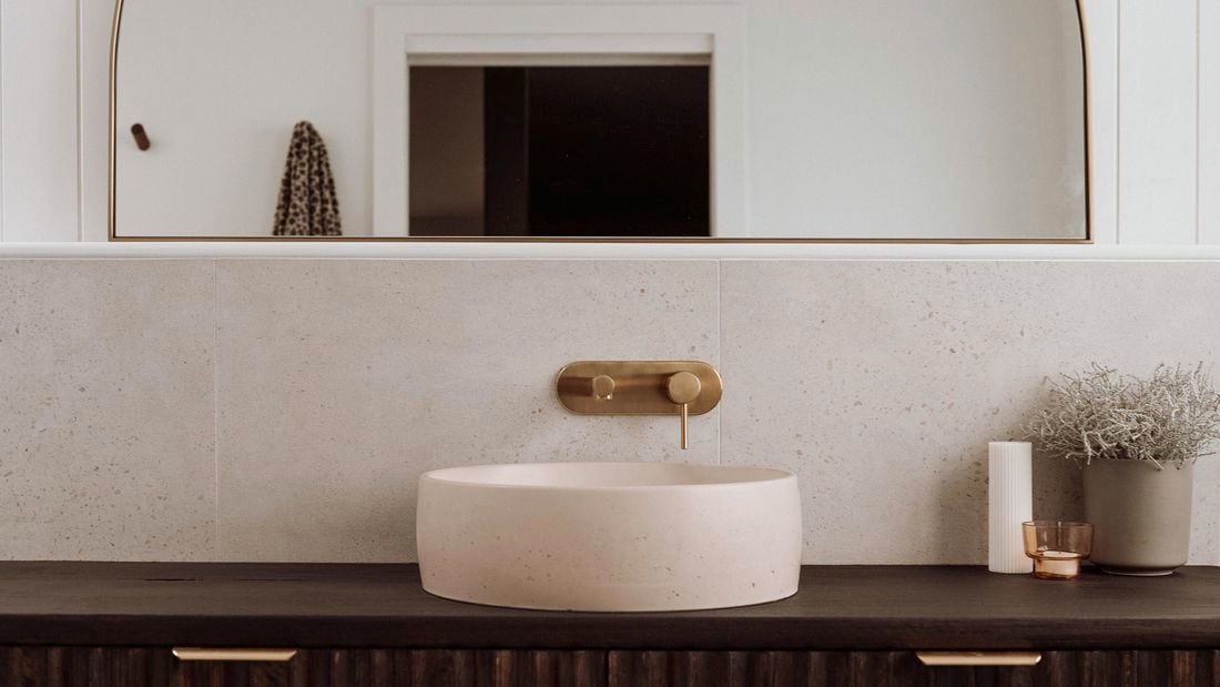 Dark brown cabinet and light pink sink with a gold tap and a semicircular mirror.