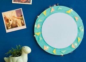 A round decorative wall mirror with a teal painted frame, adorned with pastel triangle embellishments and string lights, hanging on a blue wall next to two polaroid photos and a dinosaur-shaped plant pot