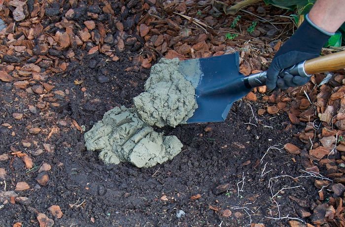 A person wearing gloves uses a shovel to place wet concrete into a hole in the soil surrounded by mulch.