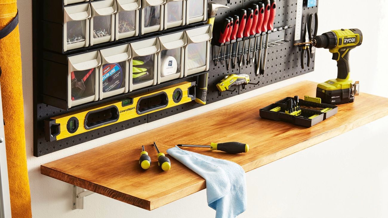 Wall‑mounted pegboard with small‑parts bins and hand tools above a timber workbench; a cordless drill, bit set, screwdrivers, spirit level and a cloth on the bench