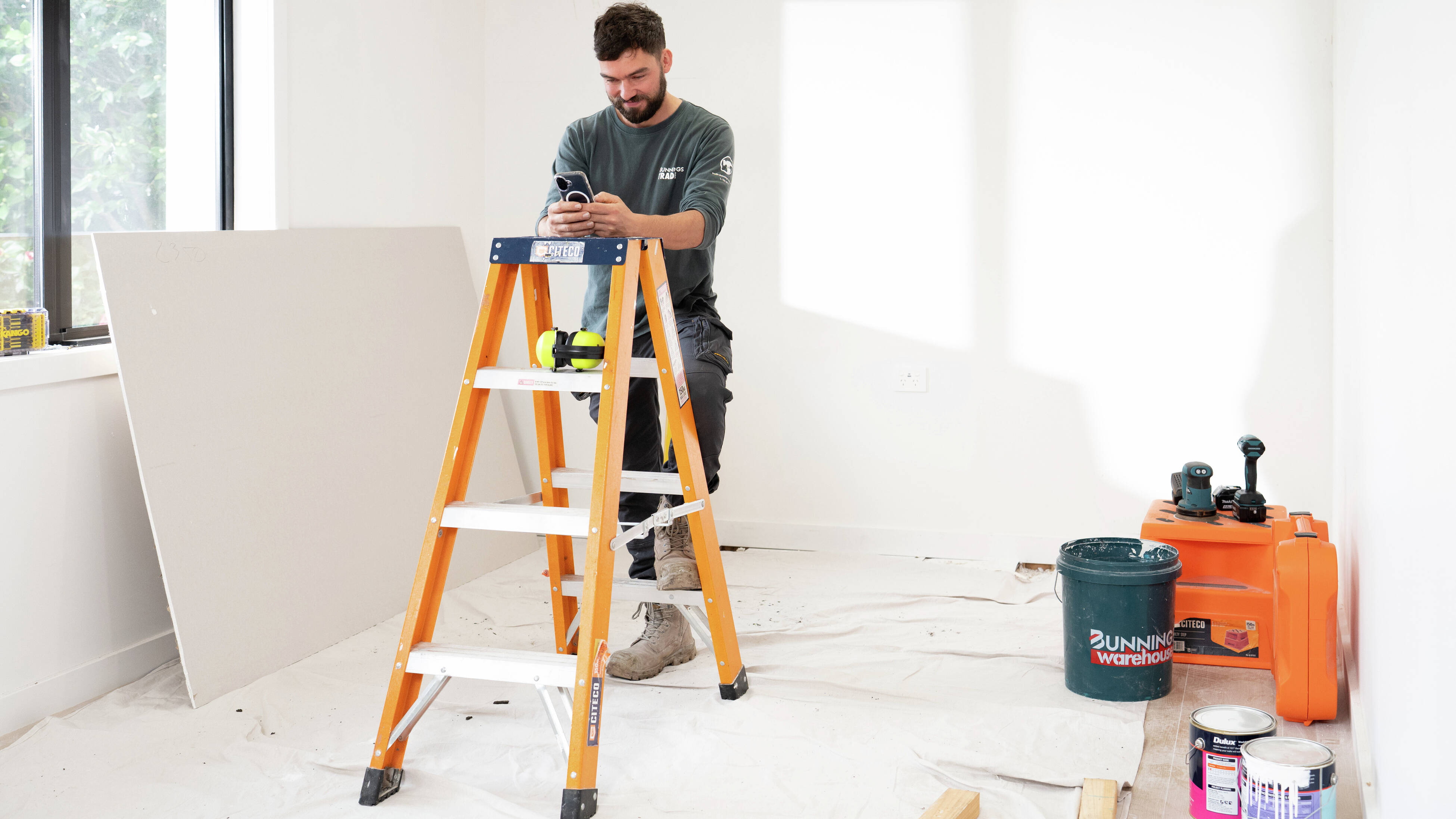 A man in a Bunnings Trade long-sleeved shirt is looking at his phone while leaning on a Citeco ladder on a jobsite featuring a Bunnings bucket, paint tins and power tools.