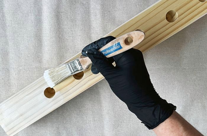 A hand wearing a black glove applies white paint using a paintbrush to a piece of timber with holes drilled into it with a drop sheet underneath.