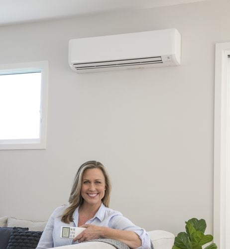 Woman sitting on couch holding air conditioner remote with air conditioning unit on wall behind her