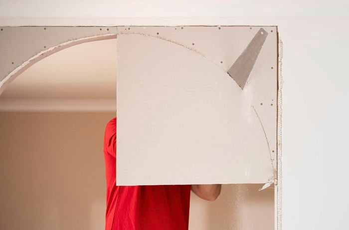 A person in a red shirt is cutting an arch shape from a plasterboard using a hand saw.
