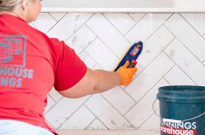 A Bunnings Team Member is applying grout to a tiled wall in a herringbone pattern using a grout float.