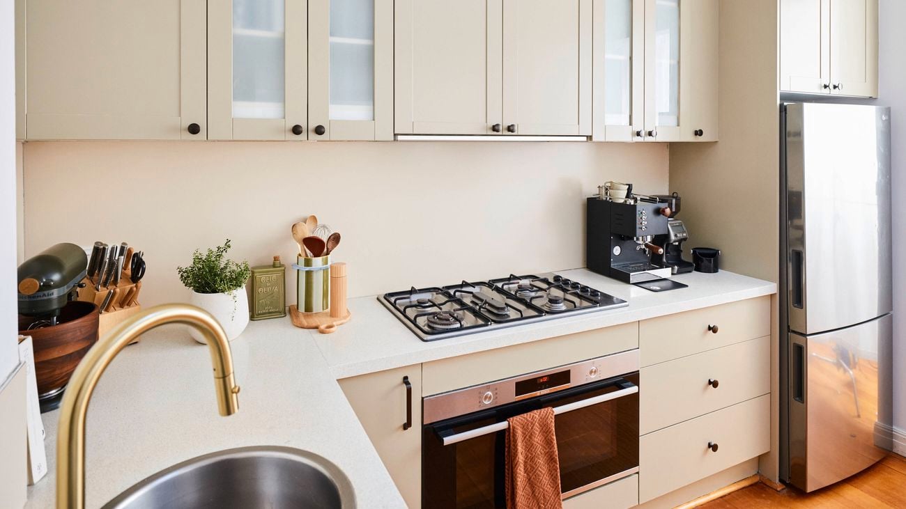 Close-up of a modern kitchen setup with beige cabinetry and white benchtops, featuring a gas cooktop above a built-in oven, a stainless steel refrigerator, and small appliances including a coffee machine, with a sink and brass tap visible in the foreground.