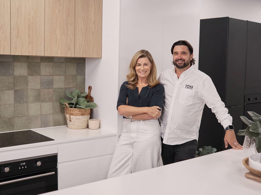 TV’s Deborah Hutton and Adam Smith of Toki Construction standing in a modern kitchen with white countertops, light timber overhead cabinets, dark grey or black lower cabinets and appliances, and a tiled splashback