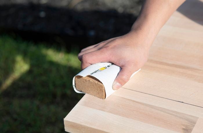 The surface of a desk being sanded with sandpaper wrapped around a scrap piece of timber