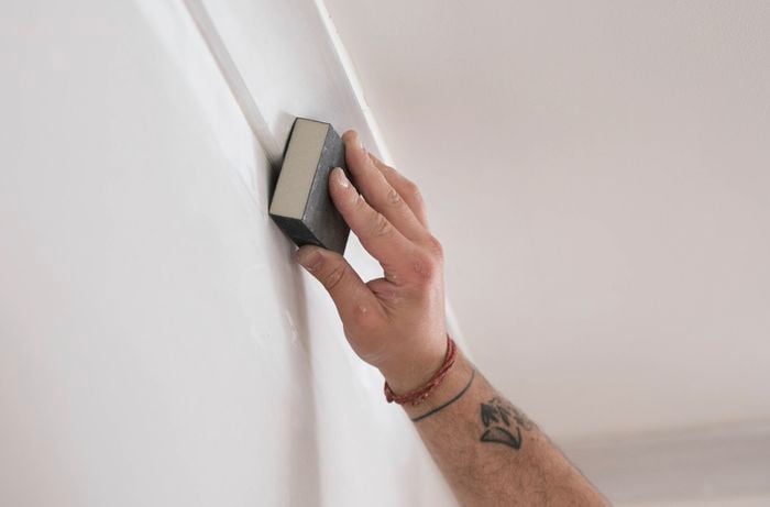 A person is using a sanding block to smooth the surface of a wall near a cornice to remove any rough patches.