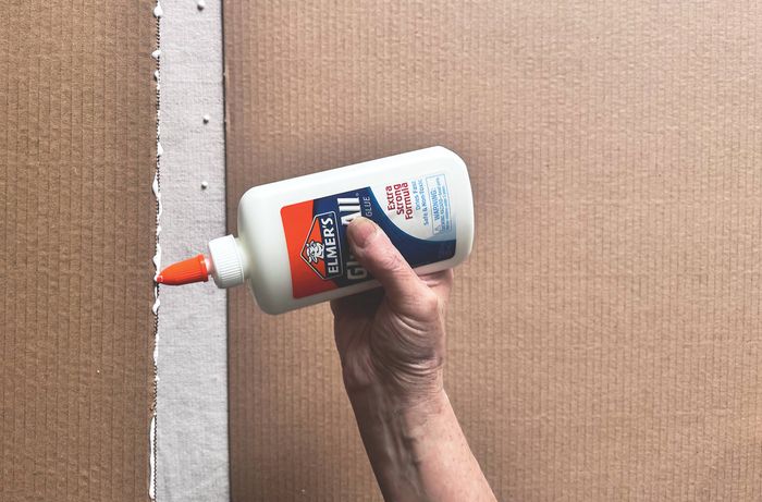 A gloved hand using a brush to paint a piece of cardboard white between strips of blue masking tape.