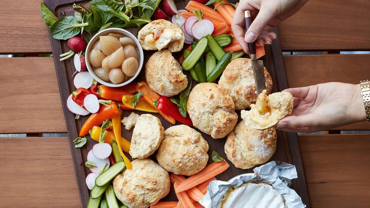 A close-up image of two hands putting brie cheese on a saltbush damper roll picked up from a brown timber tray filled with a variety of vegetables and saltbush damper rolls