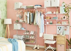 A bedroom with pink and green wallpaper, showing the corner of a bed and a floor lamp and open shelving on one wall. The shelving has different sections for hanging clothes,  shoe storage, cube storage, a small wooden desk and chair and stationery.