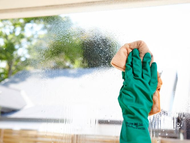 Person wearing a green rubber glove cleaning a glass window with an orange cloth, with blurred trees and buildings visible in the background.