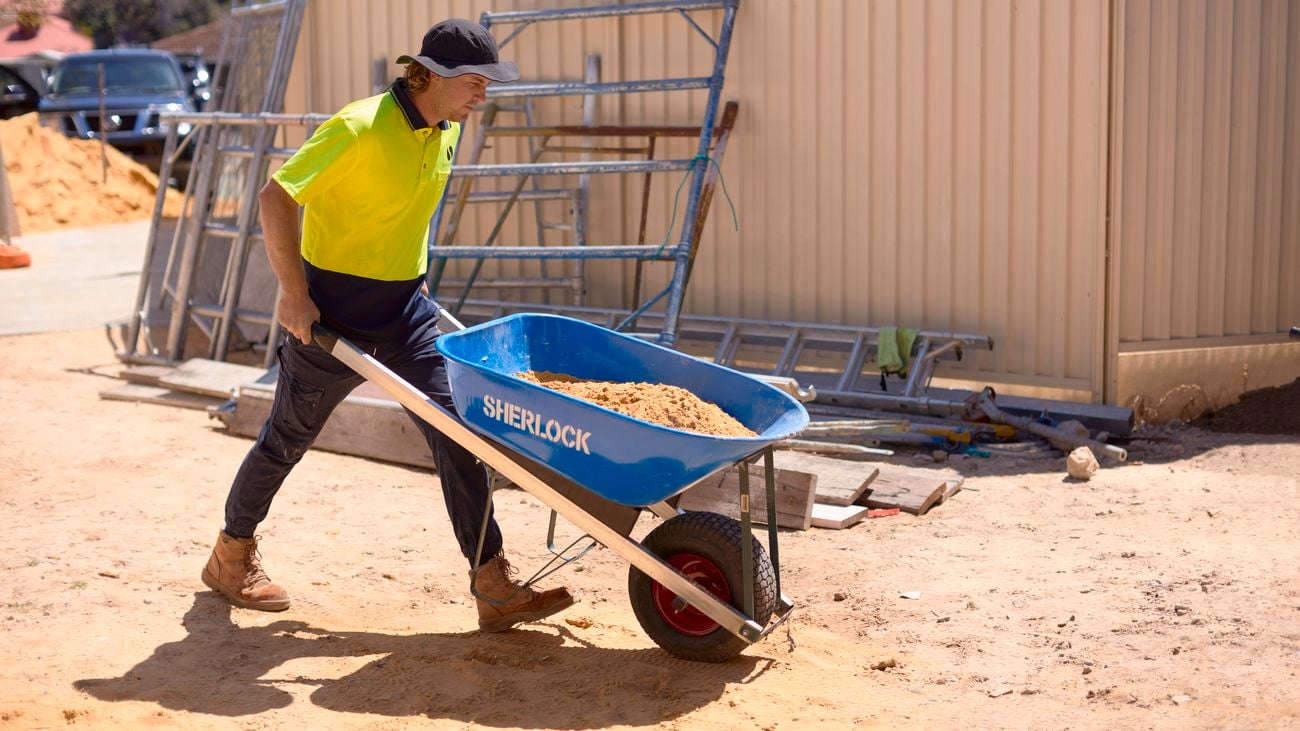 A tradesperson wearing a bucket hat, yellow hi-vis shirt and black pants pushes a blue Sherlock wheelbarrow on site.