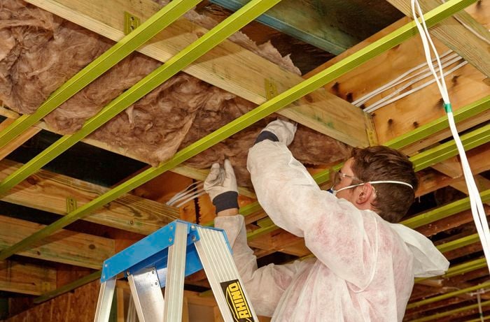 A person wearing protective gear standing on a ladder to install an insulation batt between joists in a ceiling