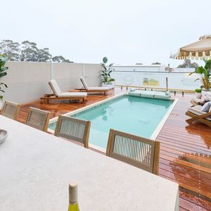 Outdoor pool and entertaining area featuring timber decking, sun loungers, tropical plants and a large dining table.