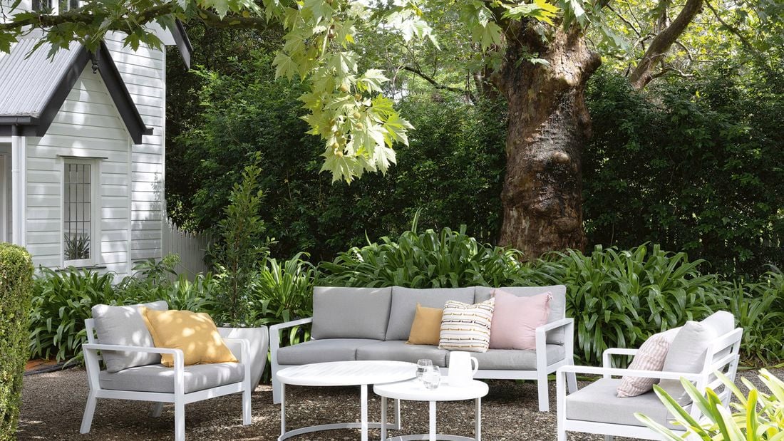  An outdoor seating area with grey and white furniture, cushions and a round table, surrounded by greenery and a large tree near a white house