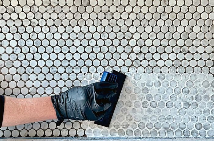 A person wearing a disposable glove uses a squeegee to grout over round mosaic tiles.