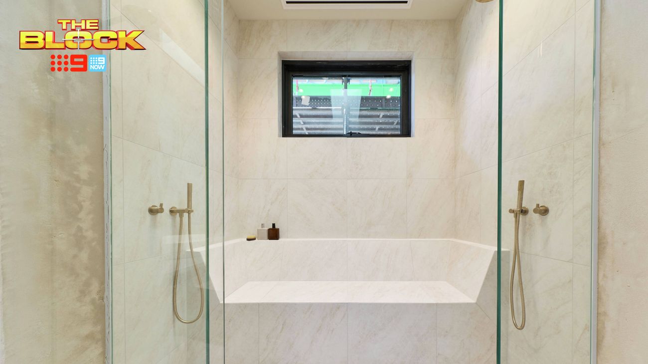 Walk-in shower with floor-to-ceiling glass panels, light stone-look tiles, built-in bench seating, brass shower fittings and a black-framed window. “The Block” logo appears in the upper-left corner.
