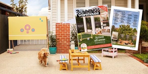 Dog carrying a toy in a family-friendly front yard play space with a tennis net, outdoor table and benches, while Bunnings Warehouse magazine cover and contents pages are displayed in the foreground.