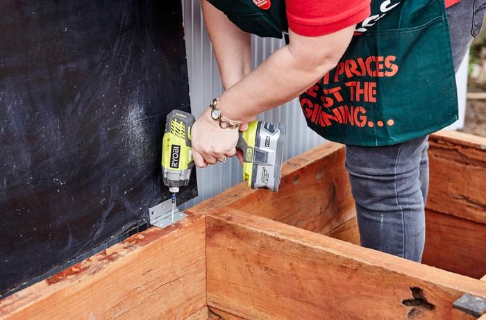 A lid hinge being screwed onto a sleeper compost bin with a power drill by a Bunnings team member