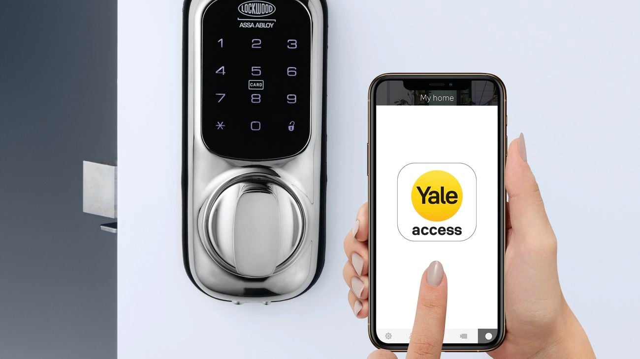 Hand holds phone with the yellow Yale app on the screen. It appears to be connected to the keypad smart door lock behind it