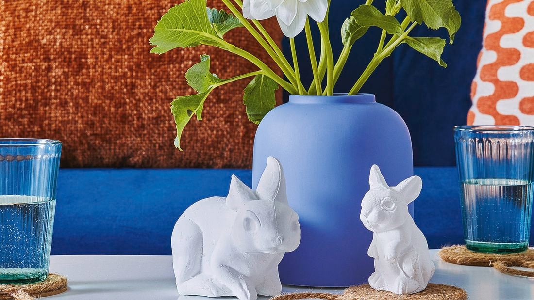 Two small bunnies made by plaster sitting in front of a blue vase with colourful cushions in the background