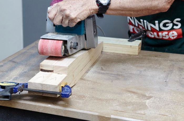 The two back supports of a wooden chair being sanded down with a belt sander, held together with a clamp