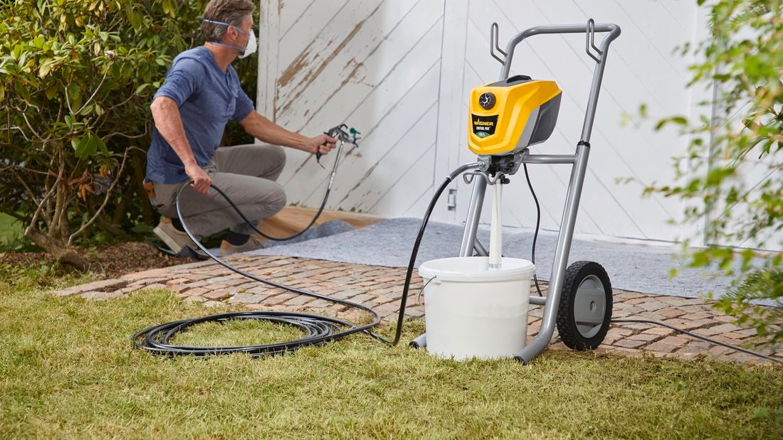 man using a spray gun to paint a shed in white