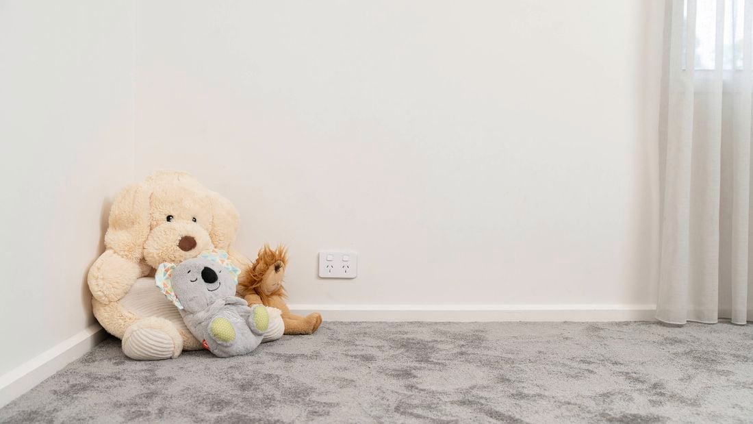 Three stuffed toys are placed on a carpeted floor next to a white wall with a power outlet nearby
