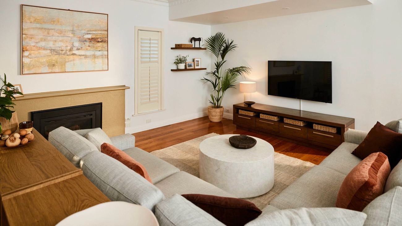 Light-filled living room with wooden floors, a wall-mounted TV above a low timber cabinet with baskets, a round coffee table on a rug, neutral sofas, indoor plants, and minimalist wall shelves.