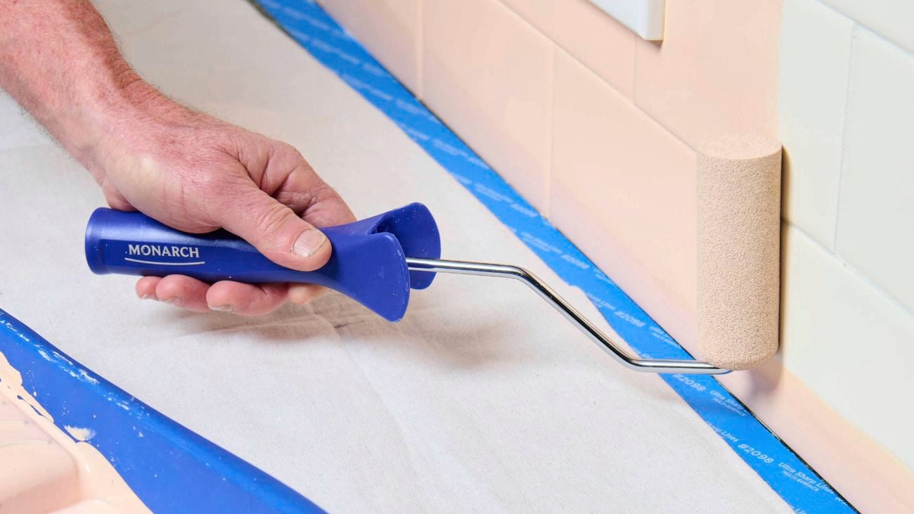 A hand holding a blue Monarch mini paint roller, painting salmon pink paint onto a tile wall
