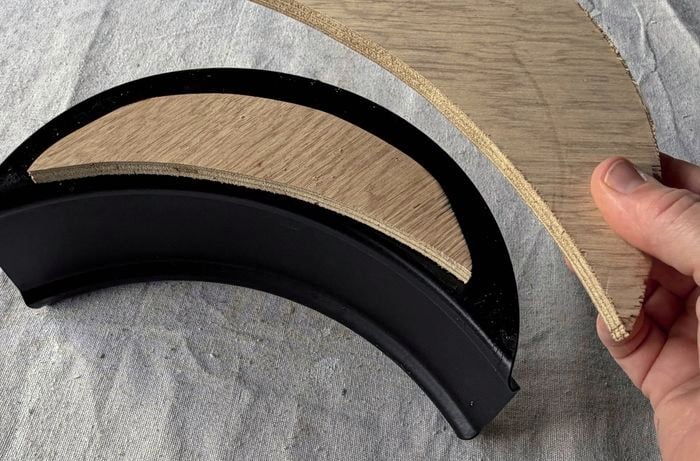 A hand holding a curved piece of plywood next to a black hose hanger with a plywood packer