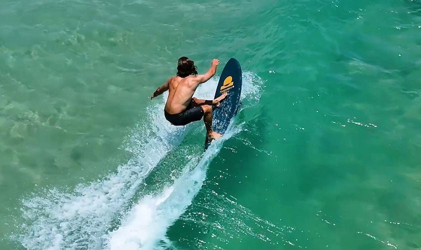Aerial view of a surfer carving a turn on a teal Summer vibes softboard in clear water.