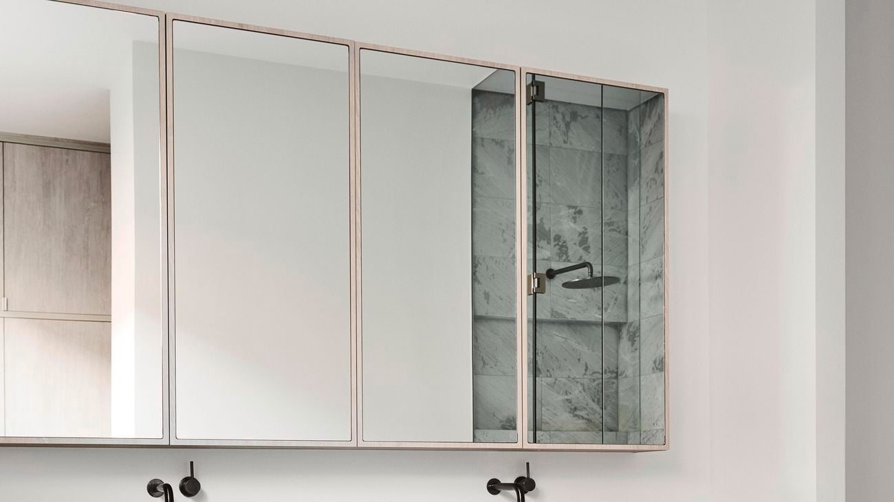 A modern white bathroom with marble countertop and mirror cabinets above reflecting the shower. A towel, a wooden table, and a bath are in view.